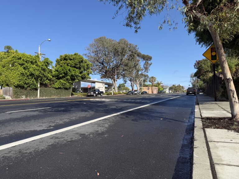 Street-level view of South Coast Highway (PCH) in Laguna Beach near the 4th Street intersection, featuring lush trees and a "Signal Ahead" asphalt marking.
