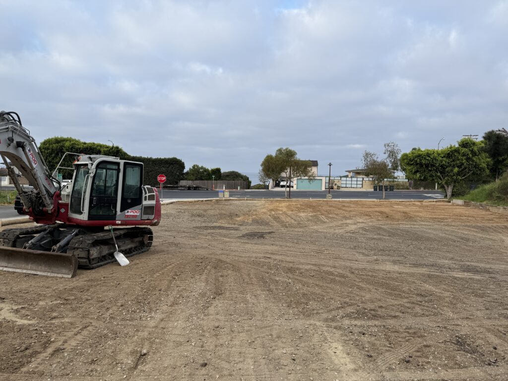 An empty dirt construction lot at the corner of PCH and 5th Ave in Laguna Beach, showing a red and white mini-excavator after the demolition of the old dentist office.
