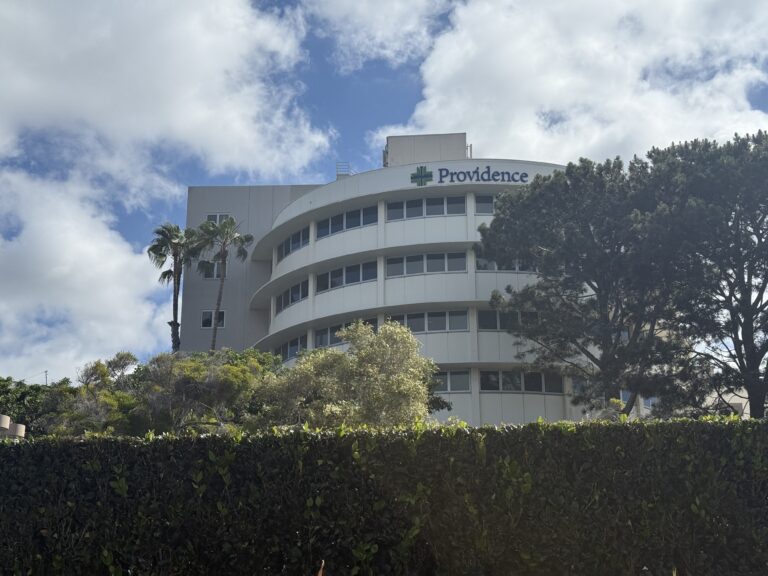 Curved white building with a Providence logo, set against a partly cloudy blue sky and surrounding greenery including palm trees and a hedge.