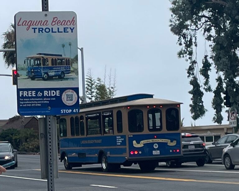 A blue and tan Laguna Beach Transit trolley driving past a street sign that reads Free to Ride Laguna Beach Trolley Stop 41.